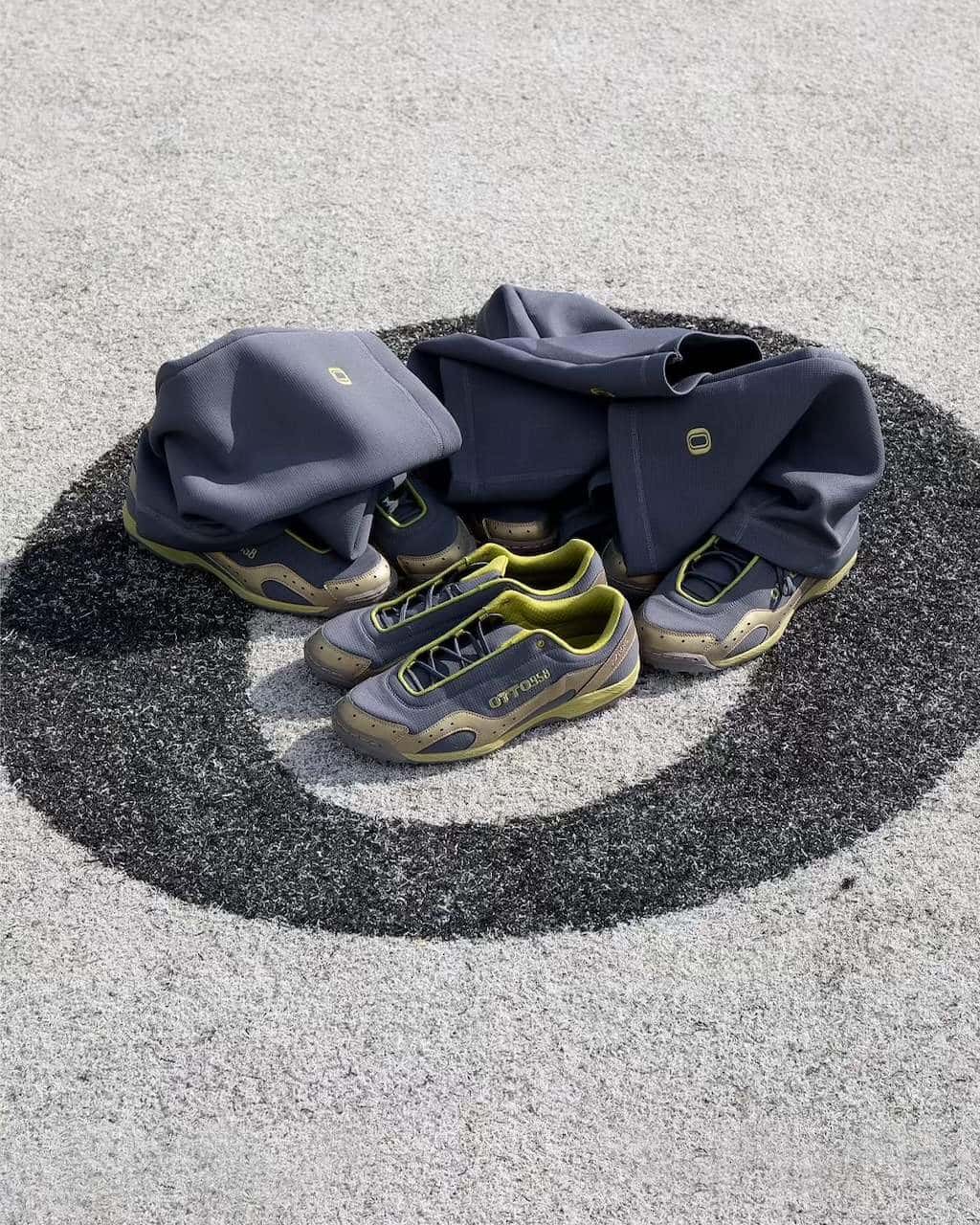 Par de ASICS GEL-BELARRAOTTO de OTTO 958 en gris oscuro con detalles verde lima y dorados, apoyado sobre un círculo pintado en el piso, con las polainas grises de ripstop desplegadas al lado